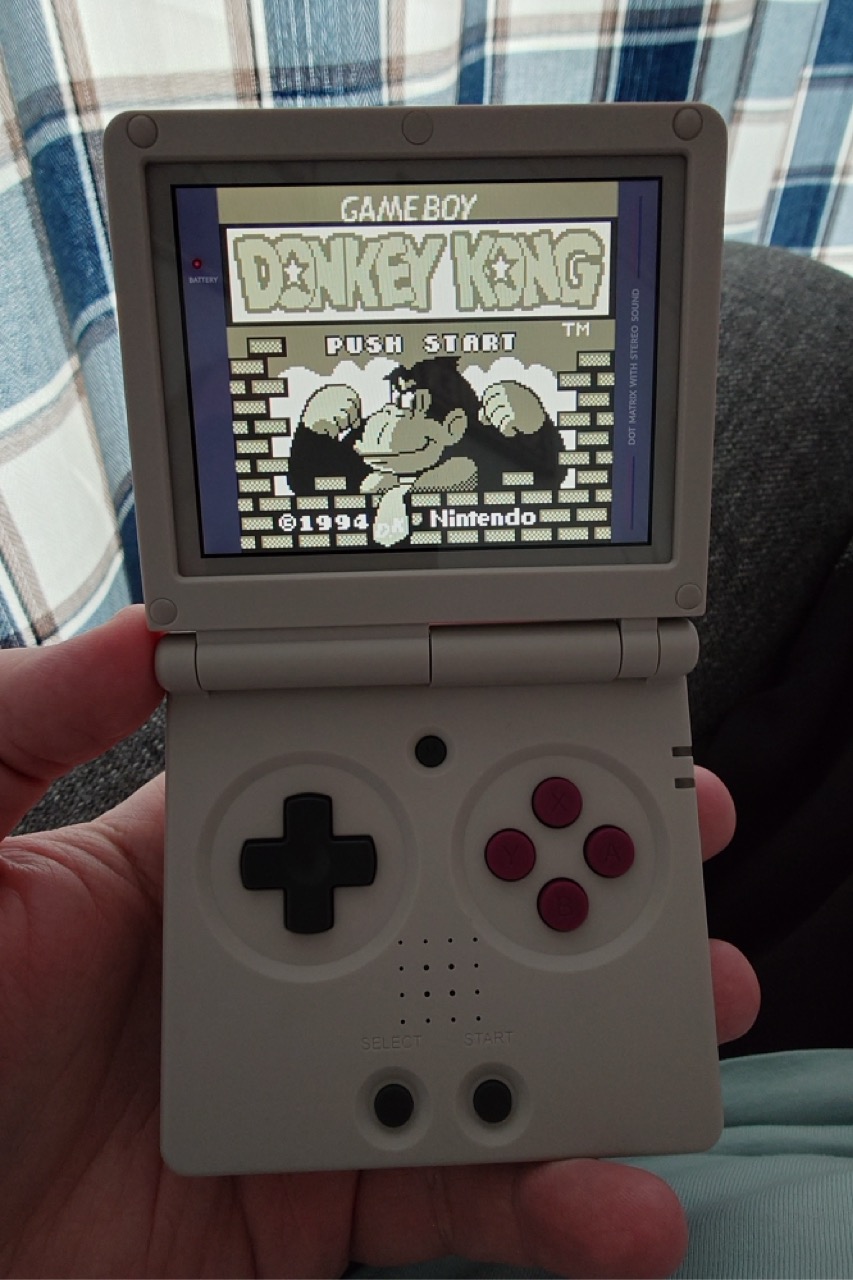 Jeg holder en ny Anbernic RG35XXSP, en sammenleggbar spillkonsoll som er modellert etter gamle Gameboy Advance SP. Konsollen er påslått og kjører Donkey Kong til Gameboy, som jeg er verdens tredje raskeste i. Gardinene i bakgrunnen og vinkelen på bildet gjør det klart at jeg sitter i godstolen min, som er det mest passende stedet for denne typen gaming